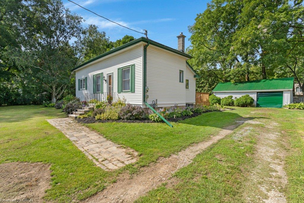 White siding exterior with green trim, stone pathway, landscaped garden beds, and a detached outbuilding with a green roof