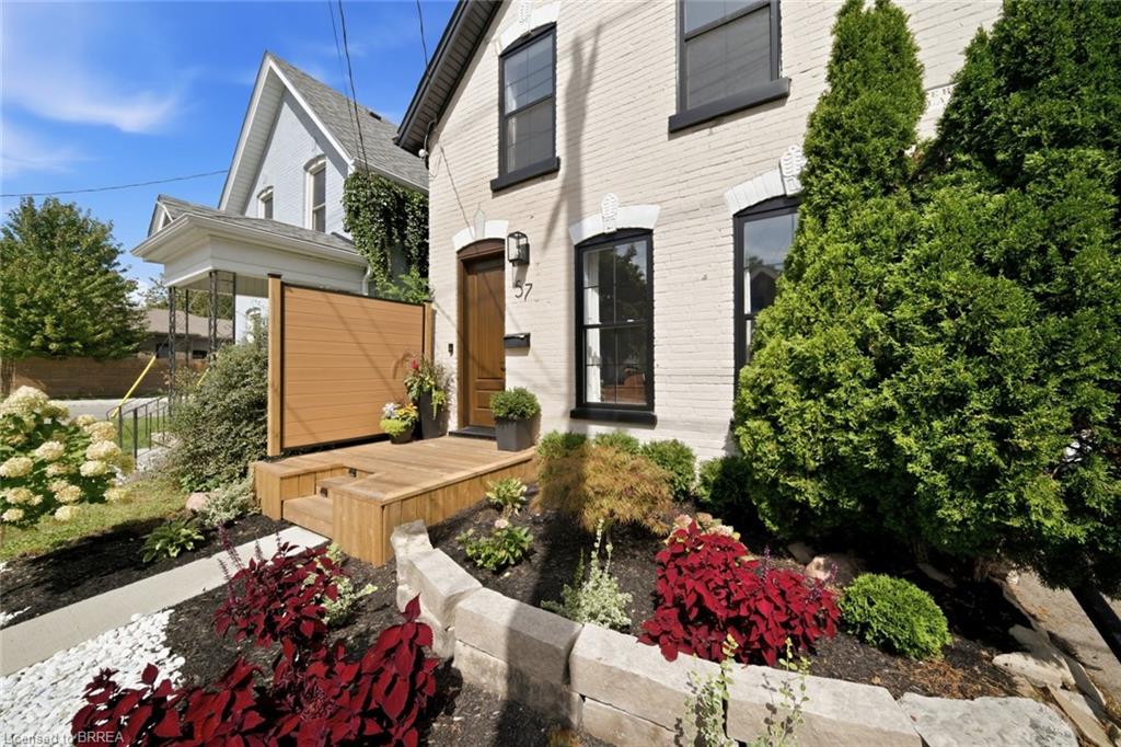 White brick exterior with black window trim, wooden entry door, elevated wooden porch, and landscaped garden beds