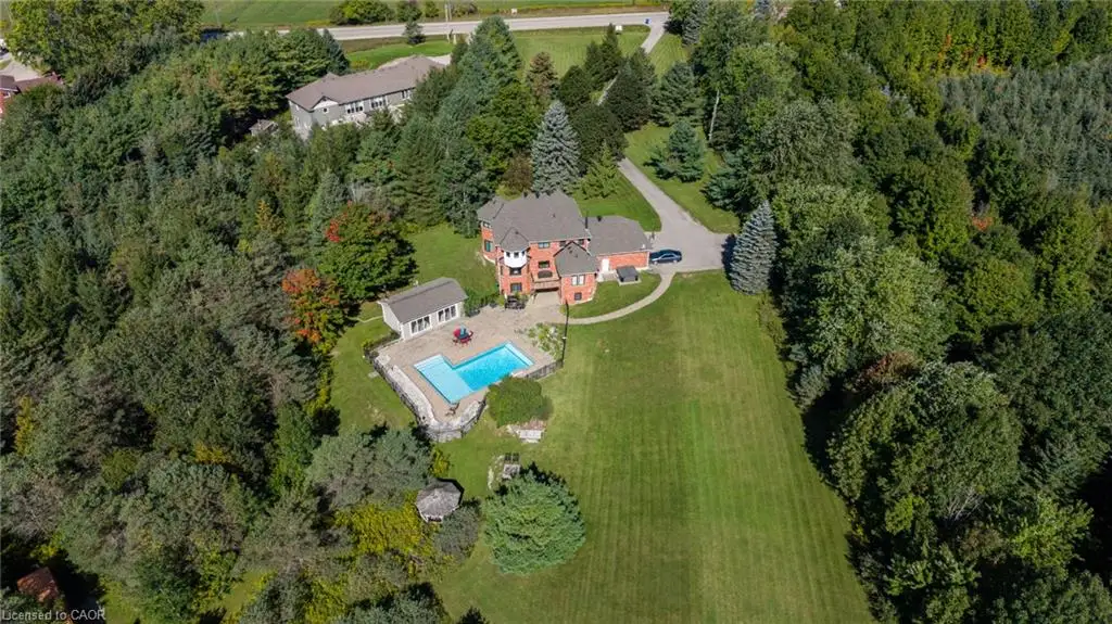 Expansive property featuring a brick exterior residence, a private swimming pool with stone surround, a dedicated pool house, a well-maintained lawn, and a surrounding wooded landscape