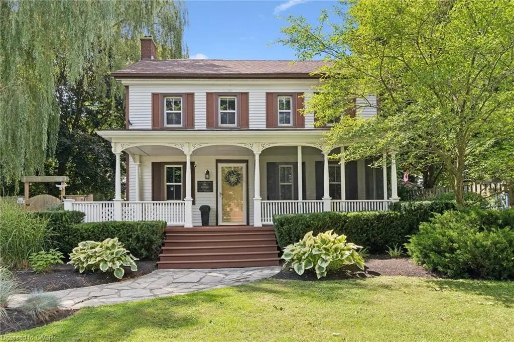 Colonial-style facade featuring white siding and brown shutters, an expansive covered porch with decorative pillars and white railing, a stone pathway, mature trees, and established landscaping