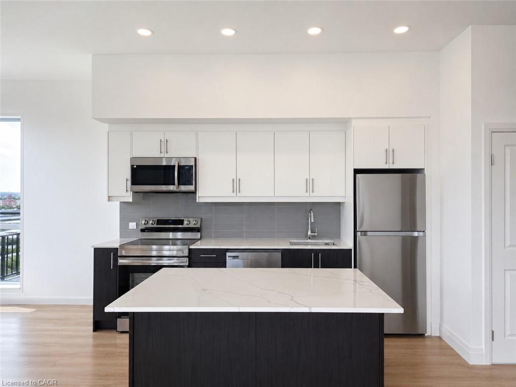 Modern kitchen featuring a central island with a light-toned countertop, stainless steel appliances, two-tone cabinetry, a gray tiled backsplash, and recessed lighting