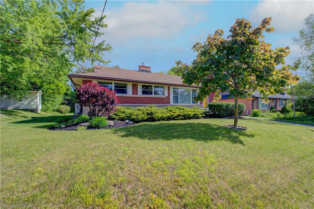Red brick exterior with white trim, a brown shingle roof, front garden beds, mature trees, and a spacious green lawn