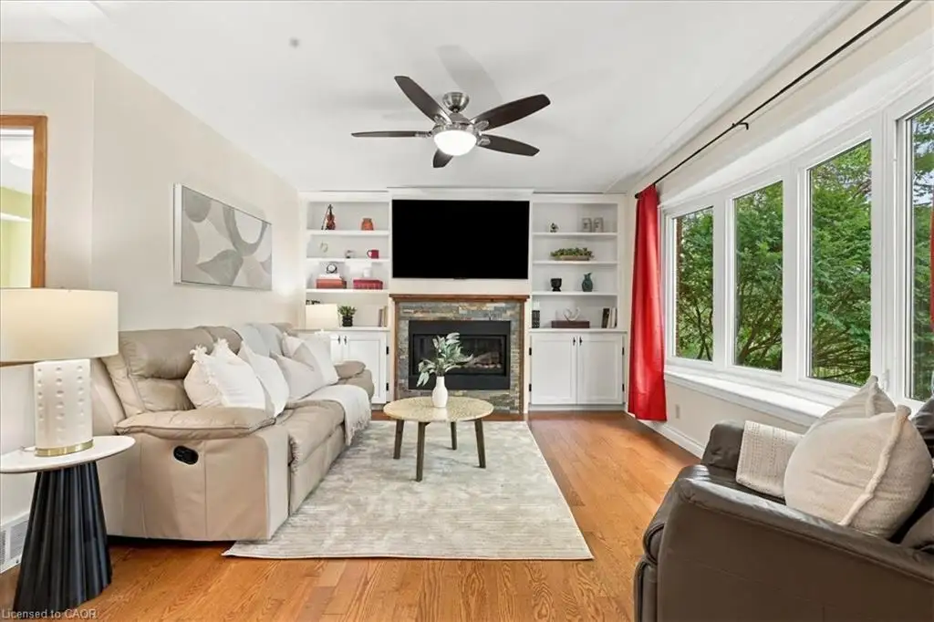 Living area featuring wood-finish flooring, a stone-surround fireplace, floor-to-ceiling built-in cabinetry, a multi-pane bay window, and a ceiling fan with integrated lighting