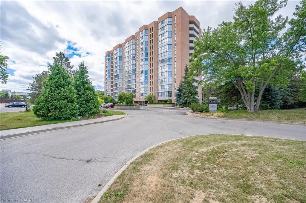 High-rise residential building featuring a red brick exterior, glass-paneled balconies, paved vehicular approach, and landscaped grounds with mature trees