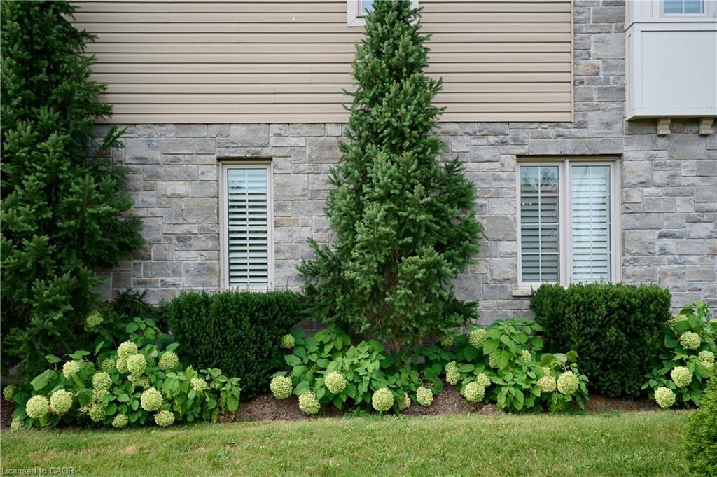 Stone facade with horizontal siding, featuring multiple windows with plantation shutters, and mature landscaping with shrubs and flowering hydrangeas