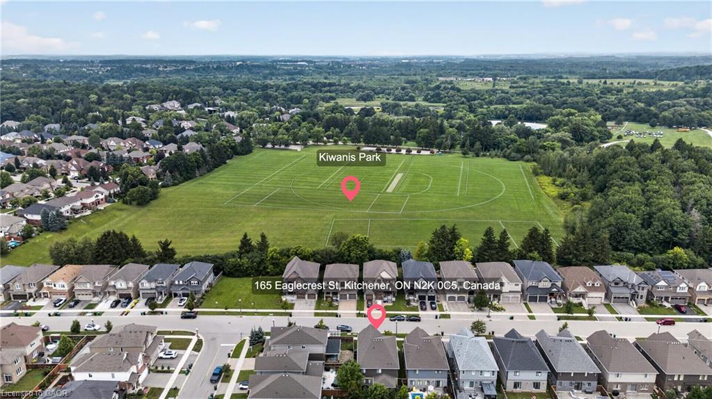 Residential street with uniform housing architecture fronting a large open field with marked sports areas and a dense tree line