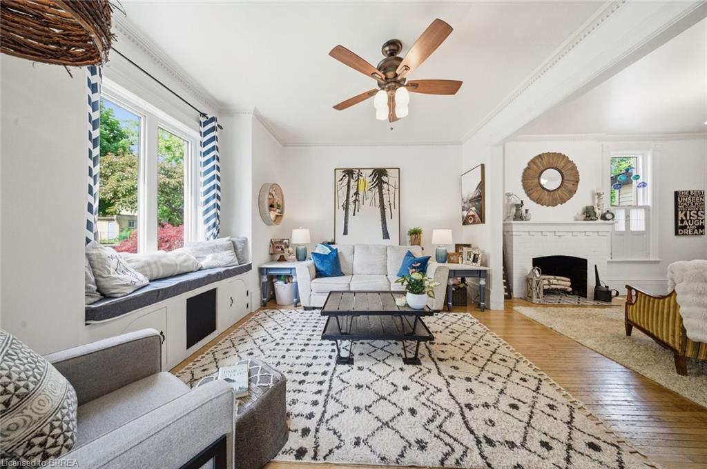 Living area featuring wood-finish flooring, a white brick fireplace, a built-in window seat with integrated storage, a ceiling fan with light fixture, and crown molding