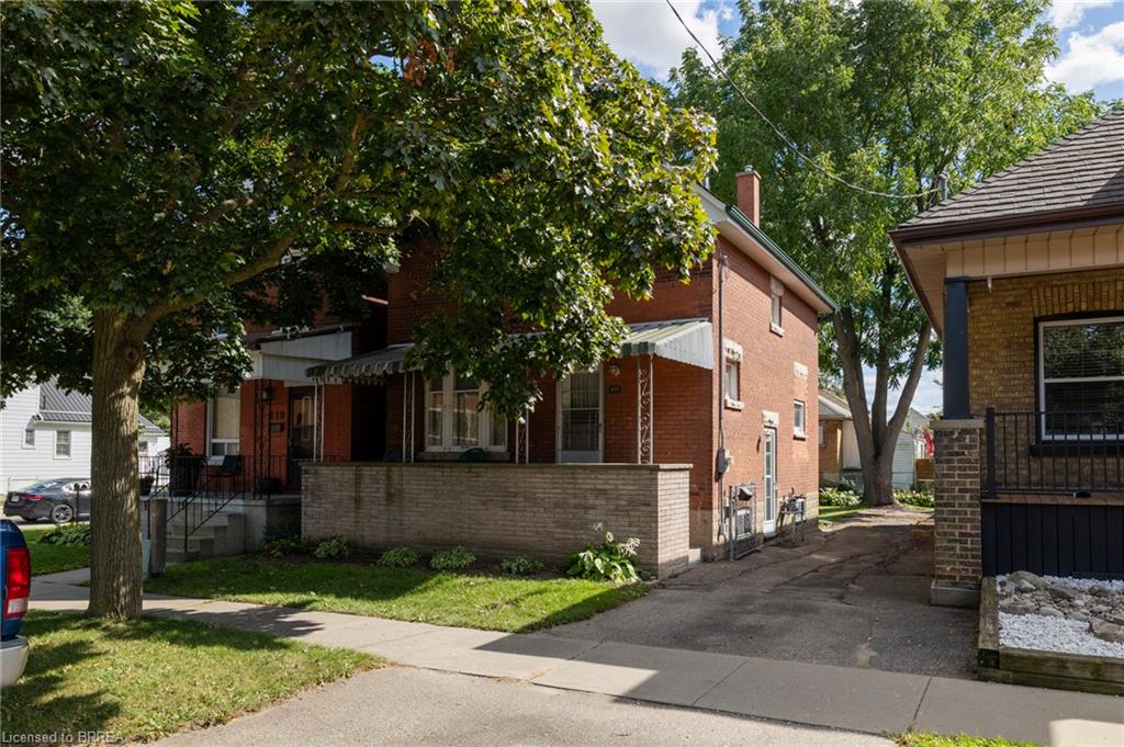 Brick exterior residence featuring a covered front porch, mature trees, a paved driveway, and a low brick privacy wall