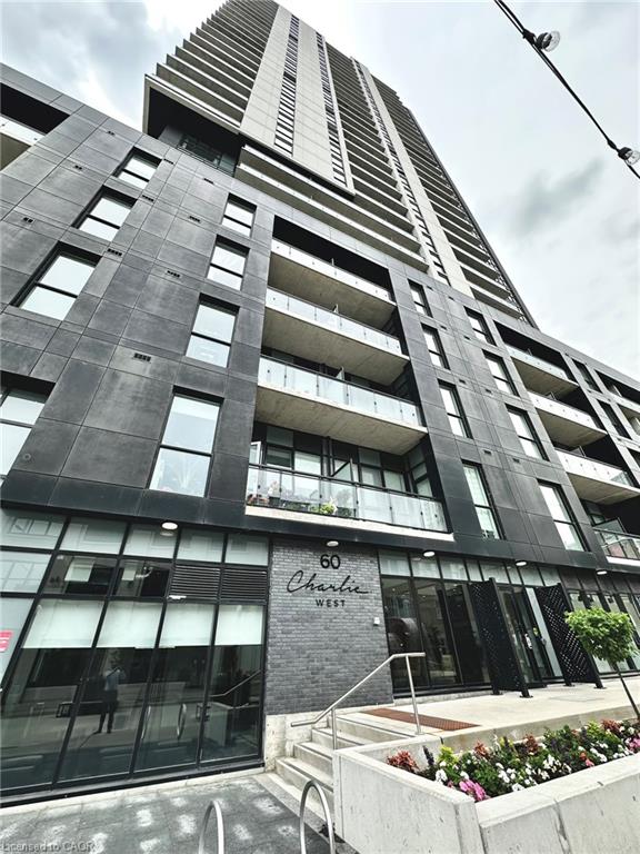 Modern high-rise building with a dark facade, numerous balconies featuring glass railings, a ground-level entrance with "Charlie West" signage, and street-level landscaping
