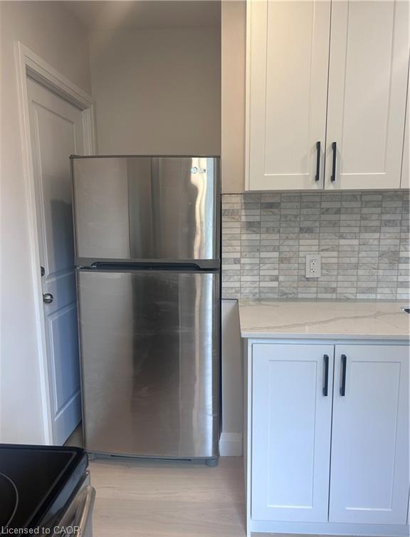 Stainless steel refrigerator, light-toned wood-finish flooring, white shaker-style cabinetry, mosaic tile backsplash, and light-toned countertop