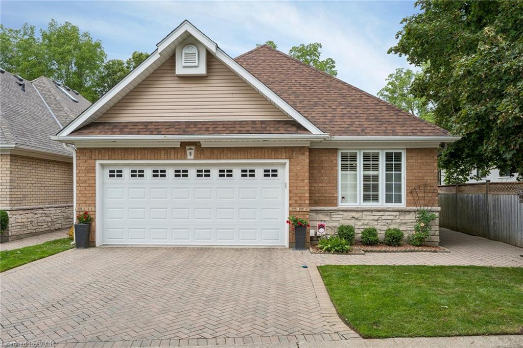 Brick and stone facade with a gable roof, featuring a paneled garage door with upper window inserts, a paver driveway, and a front window with white shutters