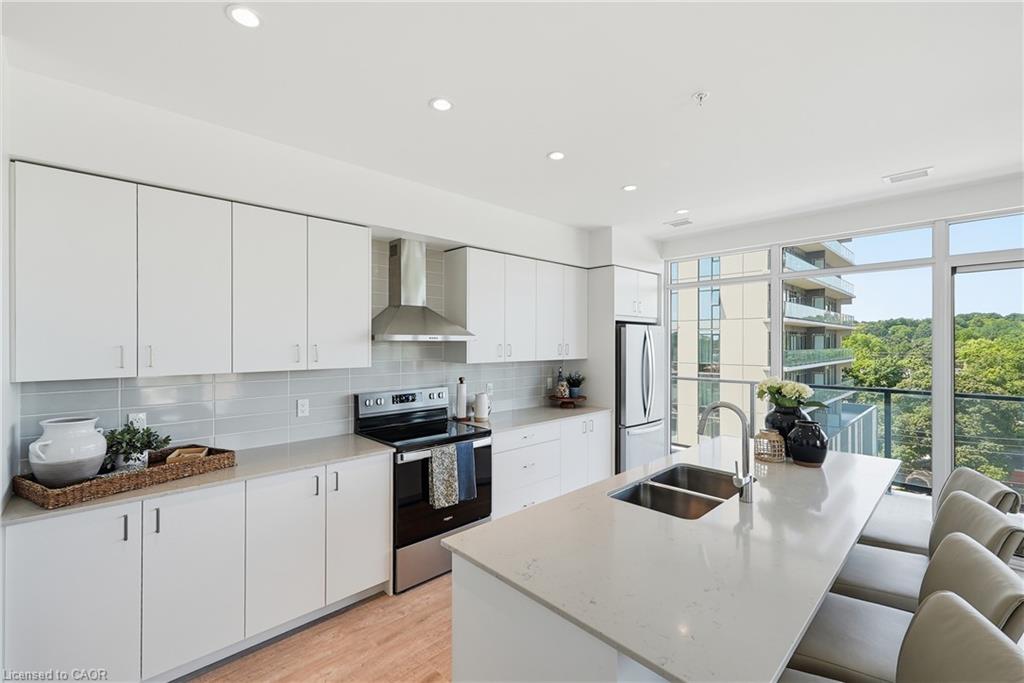 Modern kitchen featuring white cabinetry with stainless steel appliances, a quartz waterfall-edge island countertop, recessed ceiling lighting, and expansive windows overlooking tree-lined surroundings