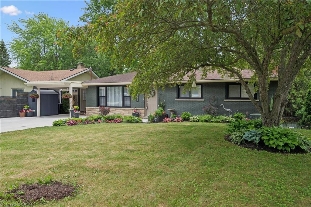 Front exterior featuring a brick facade with stone detailing, a covered carport, mature trees, manicured landscaping, and a paved driveway