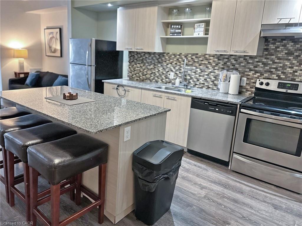 Modern kitchen featuring granite countertops, wood-finish flooring, stainless steel appliances, a multi-tone tile backsplash, and light-toned cabinetry