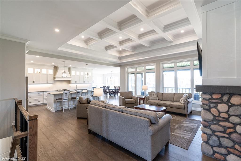 Open-concept living space with coffered ceilings, wood-finish flooring, a stone-clad fireplace, white kitchen cabinetry, and expansive windows
