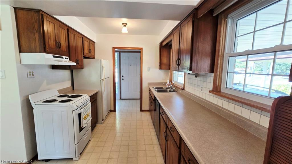 Kitchen featuring wood cabinetry, white appliances, dual-basin stainless steel sink, and light-toned countertops