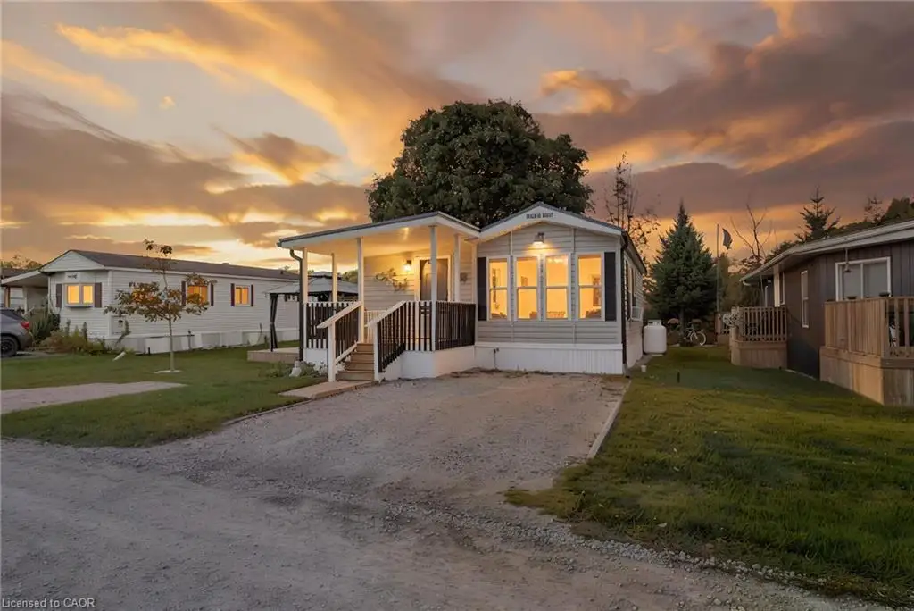 Modern manufactured home featuring a bright, open-concept living area with expansive windows, welcoming covered entry with railings, manicured lawn, and mature tree backdrop
