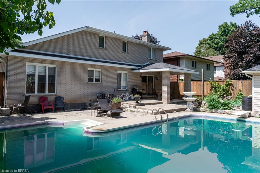 Rear exterior featuring a rectangular in-ground swimming pool with an integrated spa, a concrete pool deck, a covered patio with brick columns, and a light-colored brick facade