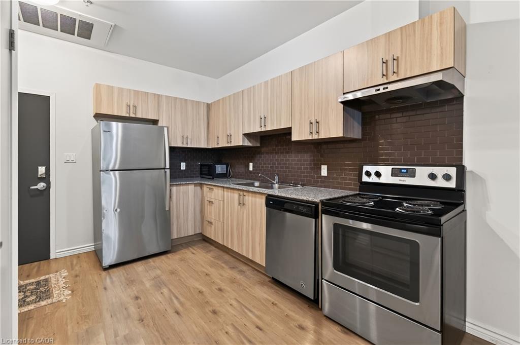 Kitchen with wood-finish flooring, light wood-finish cabinetry, stainless steel appliances, dark subway tile backsplash, and speckled countertops