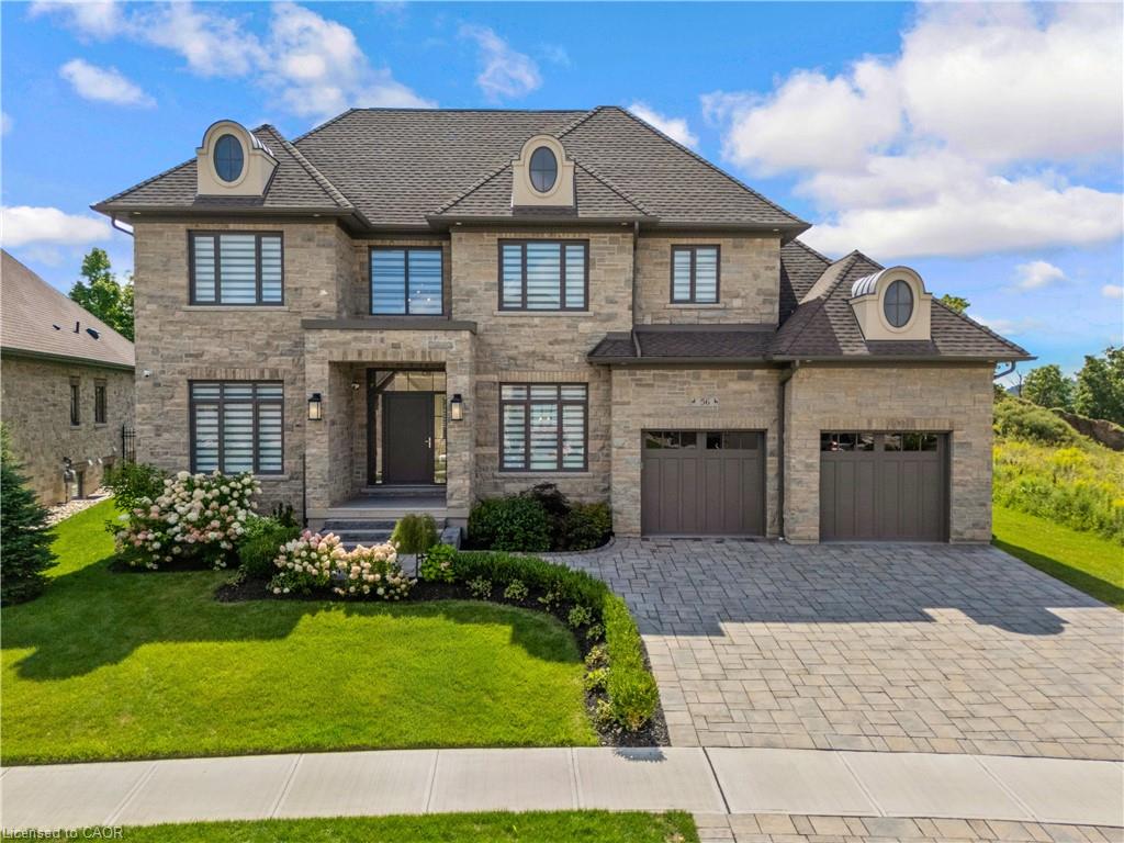 Stone facade with dormer windows, dark asphalt shingle roof, two-car garage with panel doors, paver driveway, and manicured landscaping