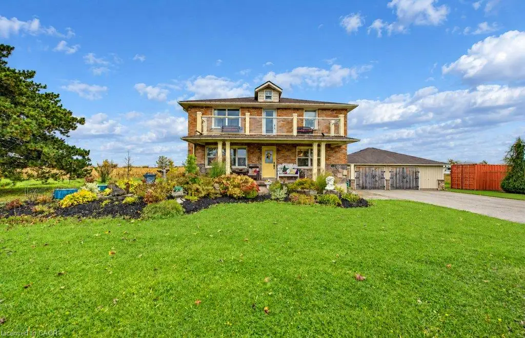 Two-story brick exterior featuring a second-level balcony, covered front porch with stone columns, detached garage with decorative wood-finish doors, and landscaped garden beds