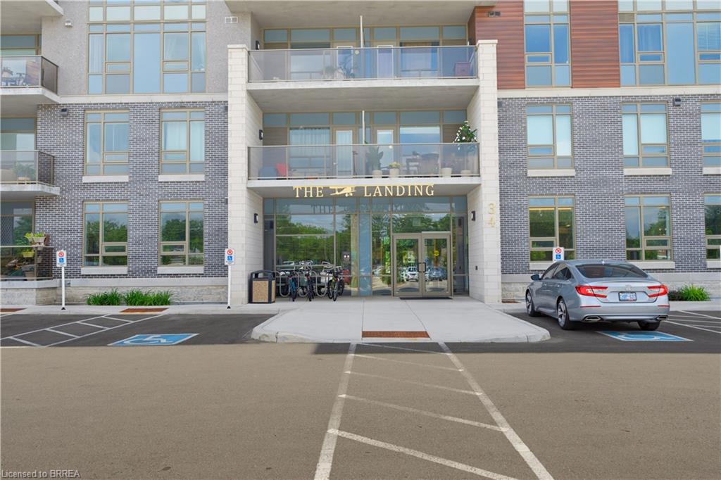 Modern building exterior featuring a prominent glass entrance, brick and stone facade, multiple balconies with glass railings, large windows, and a paved parking area