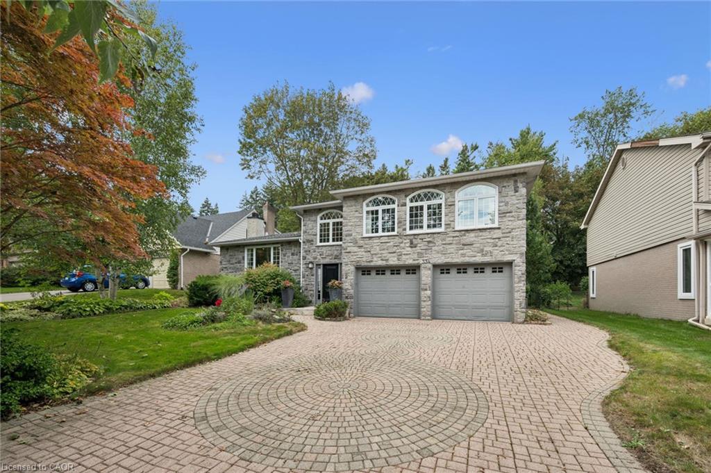 Stone facade residence featuring a paver driveway with a circular inlay, two garage doors, arched windows, and mature landscaping