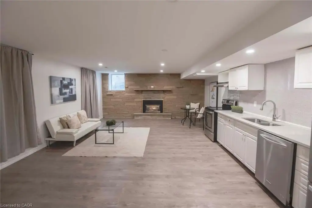 Natural stone fireplace with a wood mantel, wood-finish flooring, recessed lighting, and stainless steel kitchen appliances featuring white cabinetry