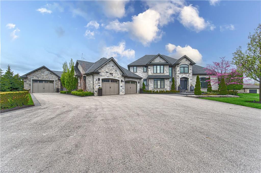 Stone exterior with multiple gables, extensive paved driveway, attached garages with panel doors, and mature landscaping