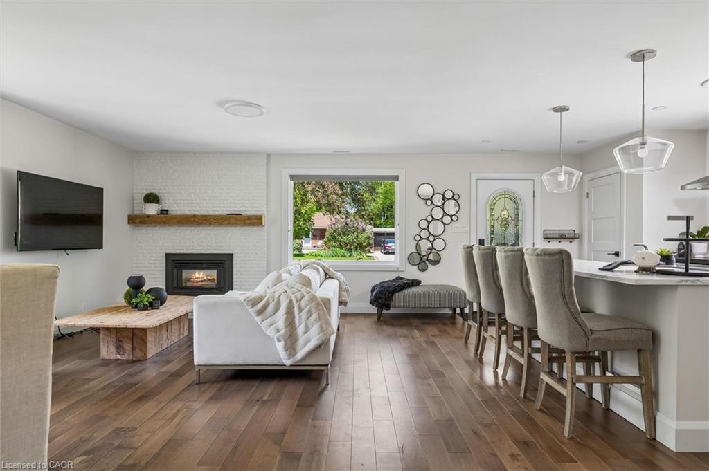 Wood-finish flooring throughout, a white brick fireplace with a wood-beam mantel, a kitchen island with seating, and clear glass pendant lighting