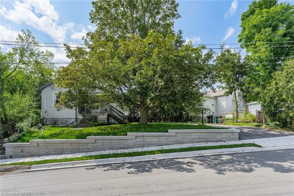 Exterior features include light-colored siding, a front porch with dark-stained railings, a mature shade tree, a landscaped retaining wall, and a paved driveway