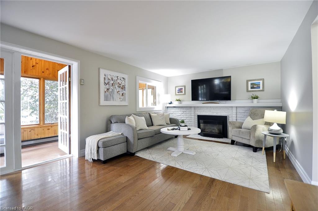 Hardwood flooring, white painted brick fireplace with electric insert, white mantel, light gray wall color, and double French doors opening to a sunroom with wood-paneled walls
