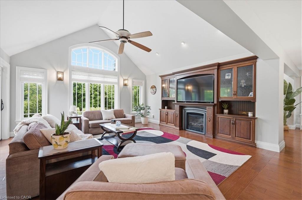 Spacious living area featuring a vaulted ceiling, abundant natural light from large windows, hand scraped engineered hardwood, a built-in entertainment center with a fireplace, and a ceiling fan
