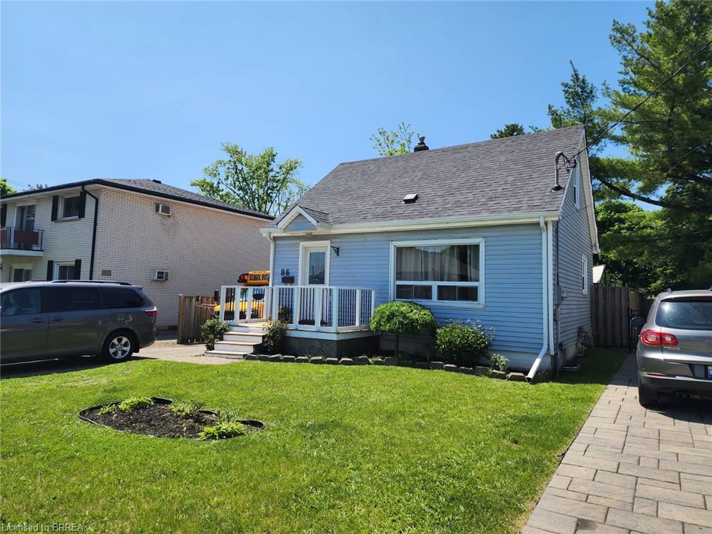 Light blue vinyl siding cottage featuring a covered front porch with white railings, peaked roofline with architectural detailing, manicured lawn with mature landscaping, paver driveway, and multi-car parking on the property