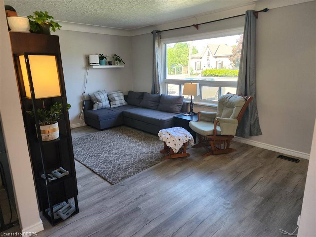 Bright and spacious living room featuring a wood-finish plank floor, large multi-pane window with natural light, contemporary sectional seating arrangement, area rug, accent shelving with decorative storage, and a modern vertical accent piece with integrated lighting