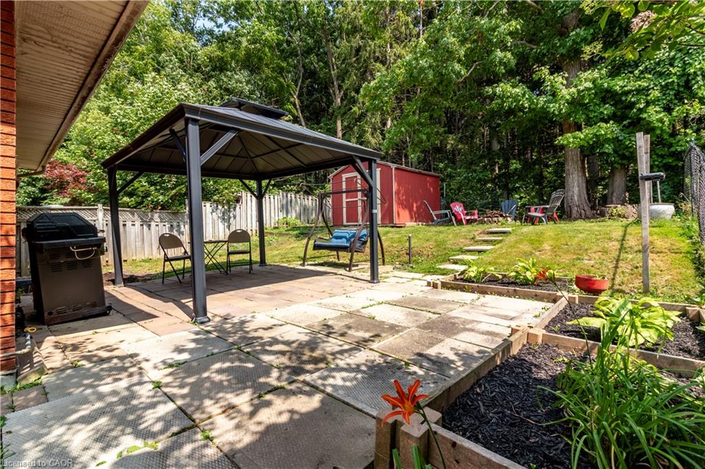 Expansive backyard featuring a paver patio with a gazebo structure, garden beds, a sloped lawn, a red storage shed, and mature trees
